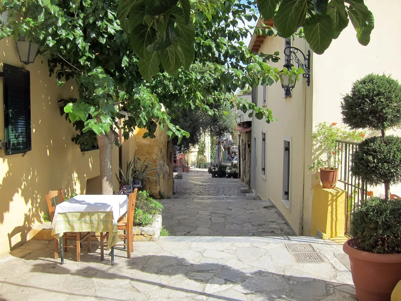A table with a checked cloth and two wooden chairs set out on a stone-paved Plaka lane under a leafy tree, between yellow-washed houses