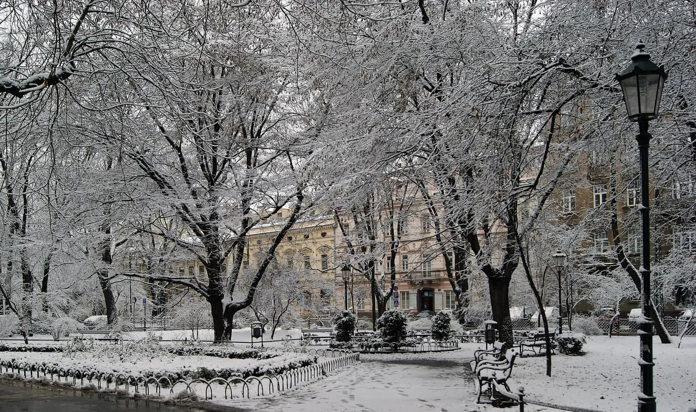 Snow-covered trees, benches and a street lamp in the Planty park, with townhouses behind