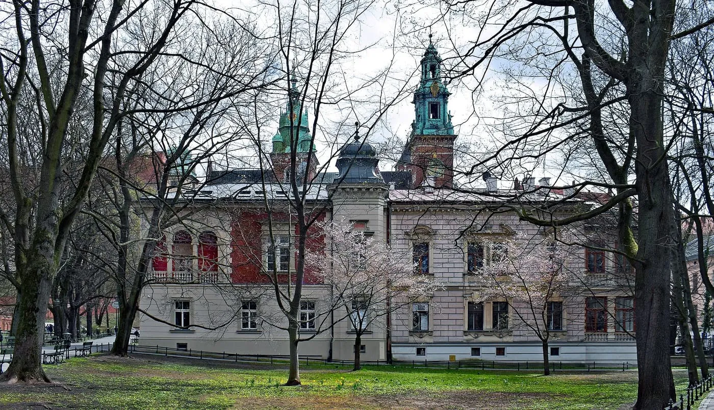 Bare trees and a cherry in blossom in the Planty park, with a villa on Straszewskiego Street and the green-roofed towers of the Wawel behind