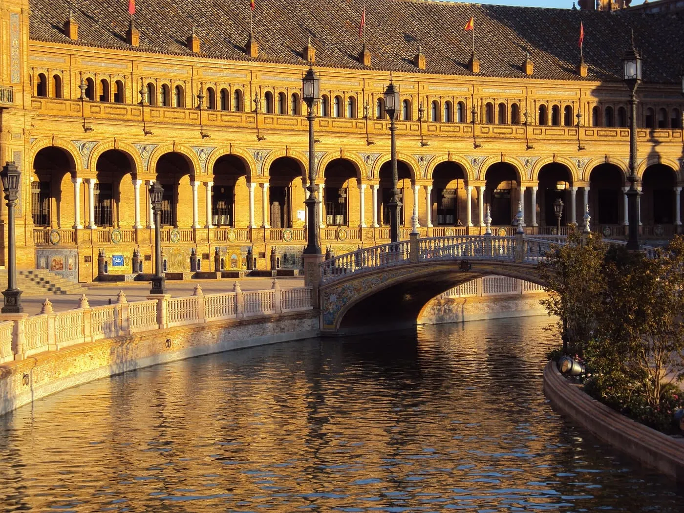 A tiled bridge over the canal of the Plaza de España, with the arcaded brick wing lit orange by low sun
