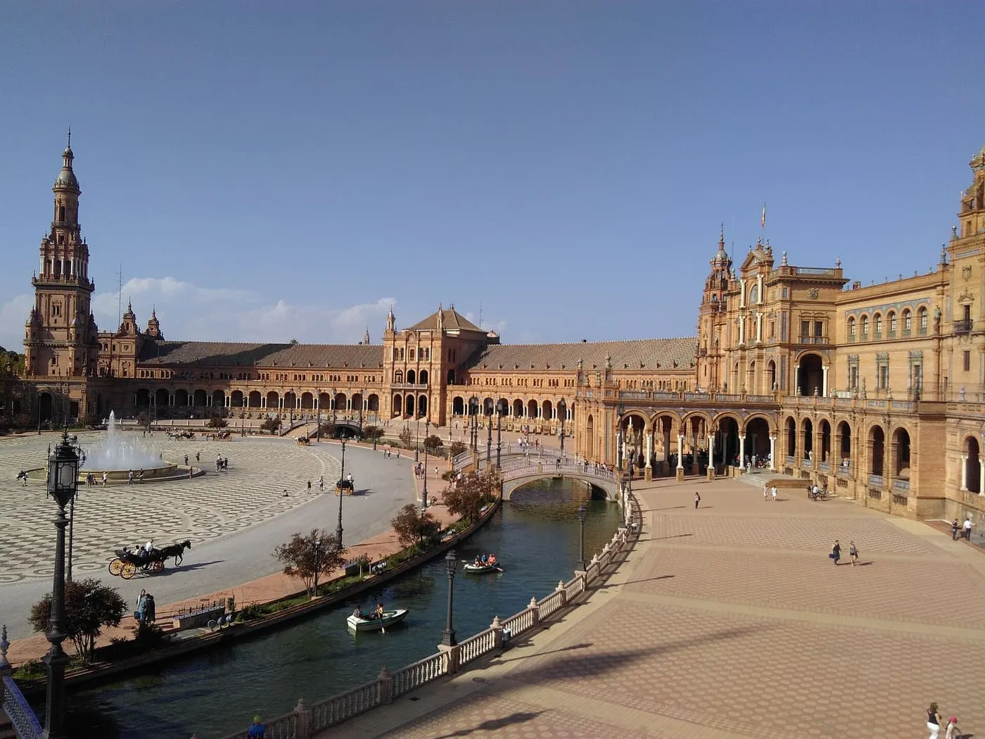The curved brick wing of the Plaza de España seen from an upper level, with the canal, a bridge, rowing boats and a horse carriage on the plaza