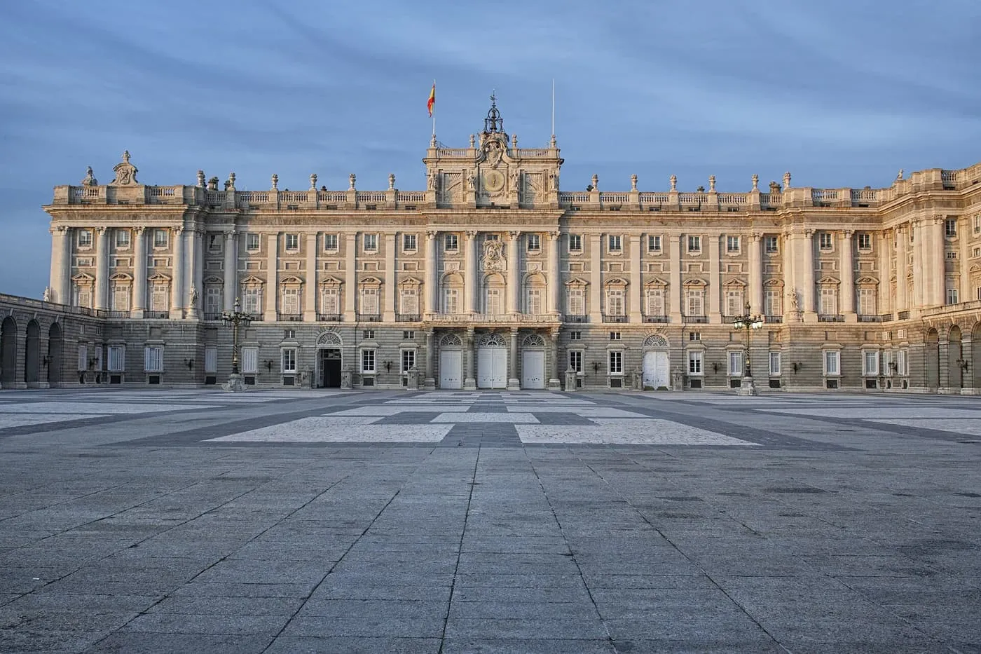 The Royal Palace of Madrid seen across the empty paved Plaza de la Armería, with its central clock, balustrade and white doors at ground level