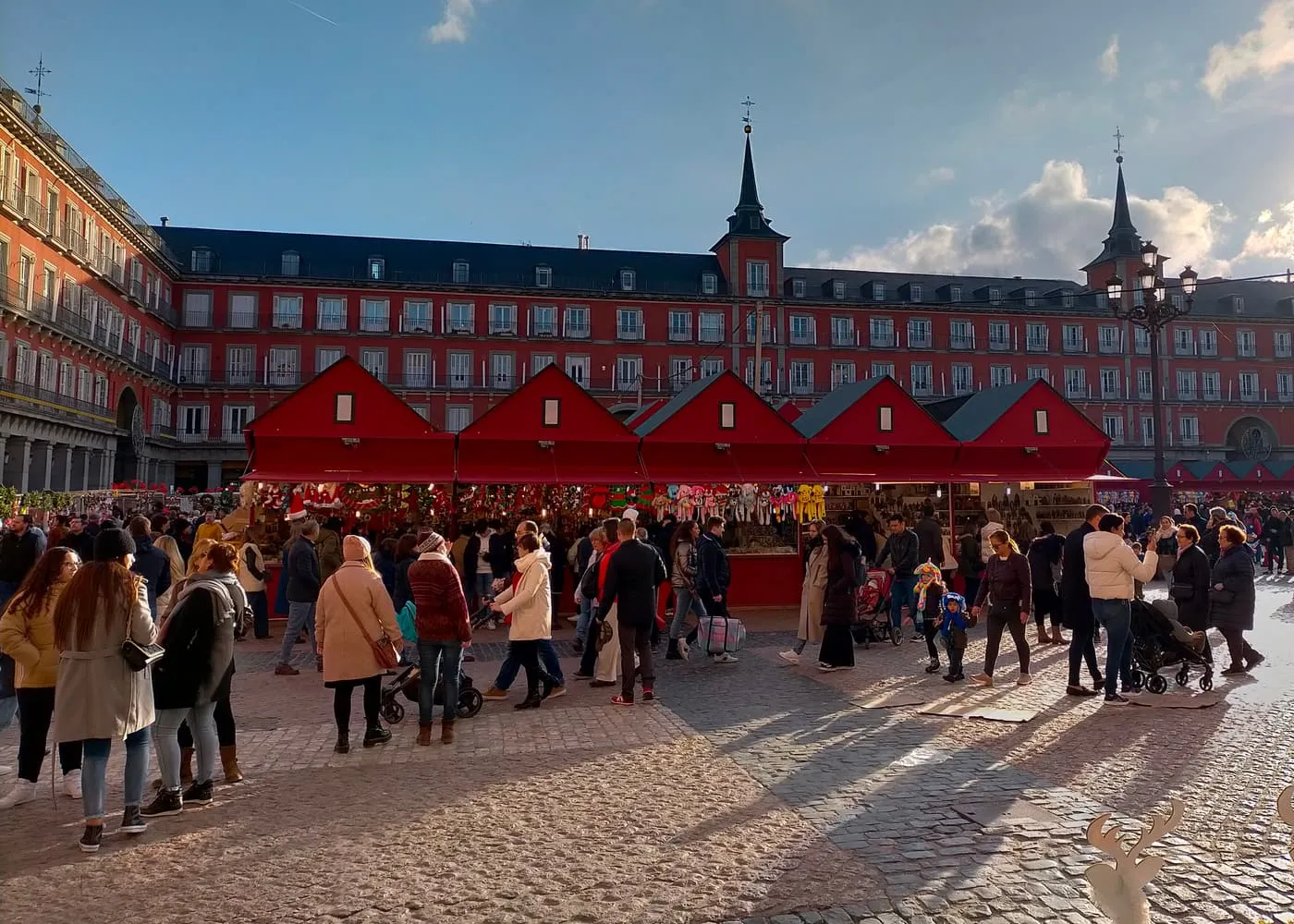 Red wooden market stalls with gabled roofs in the Plaza Mayor, with shoppers in front and the red arcaded buildings behind