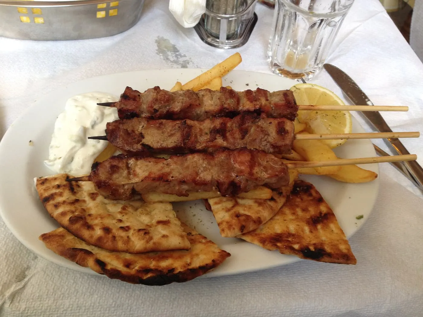 A plate of three grilled pork souvlaki skewers with a bowl of tzatziki, chips, a lemon wedge and grilled pita triangles on a paper tablecloth