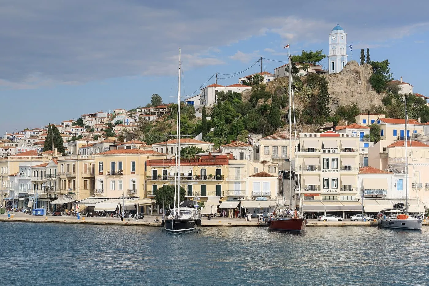 Poros's waterfront of pale neoclassical houses, with sailing yachts moored in front and the white clock tower on the rock above