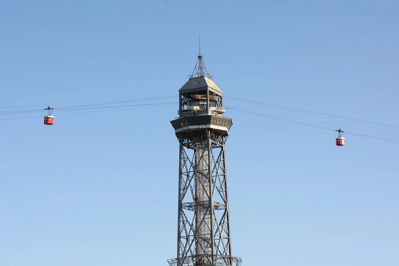 The steel lattice Torre de Jaume I with a red cabin of the port cable car on the wires to either side, against a blue sky