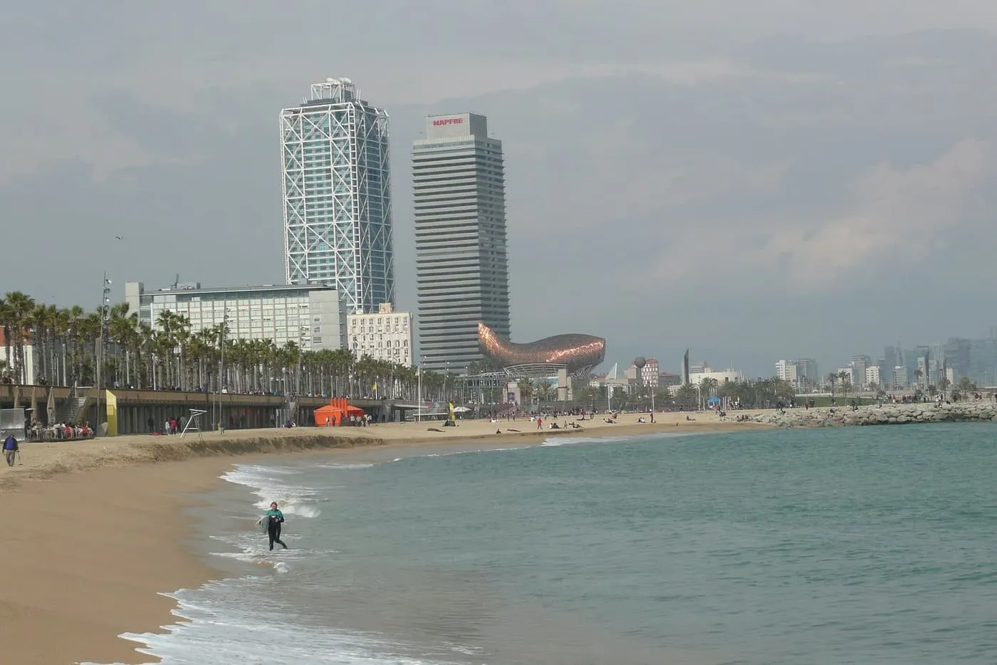 A sandy beach curving towards the two towers of the Port Olímpic, one wrapped in a white steel frame, and Frank Gehry's copper fish sculpture