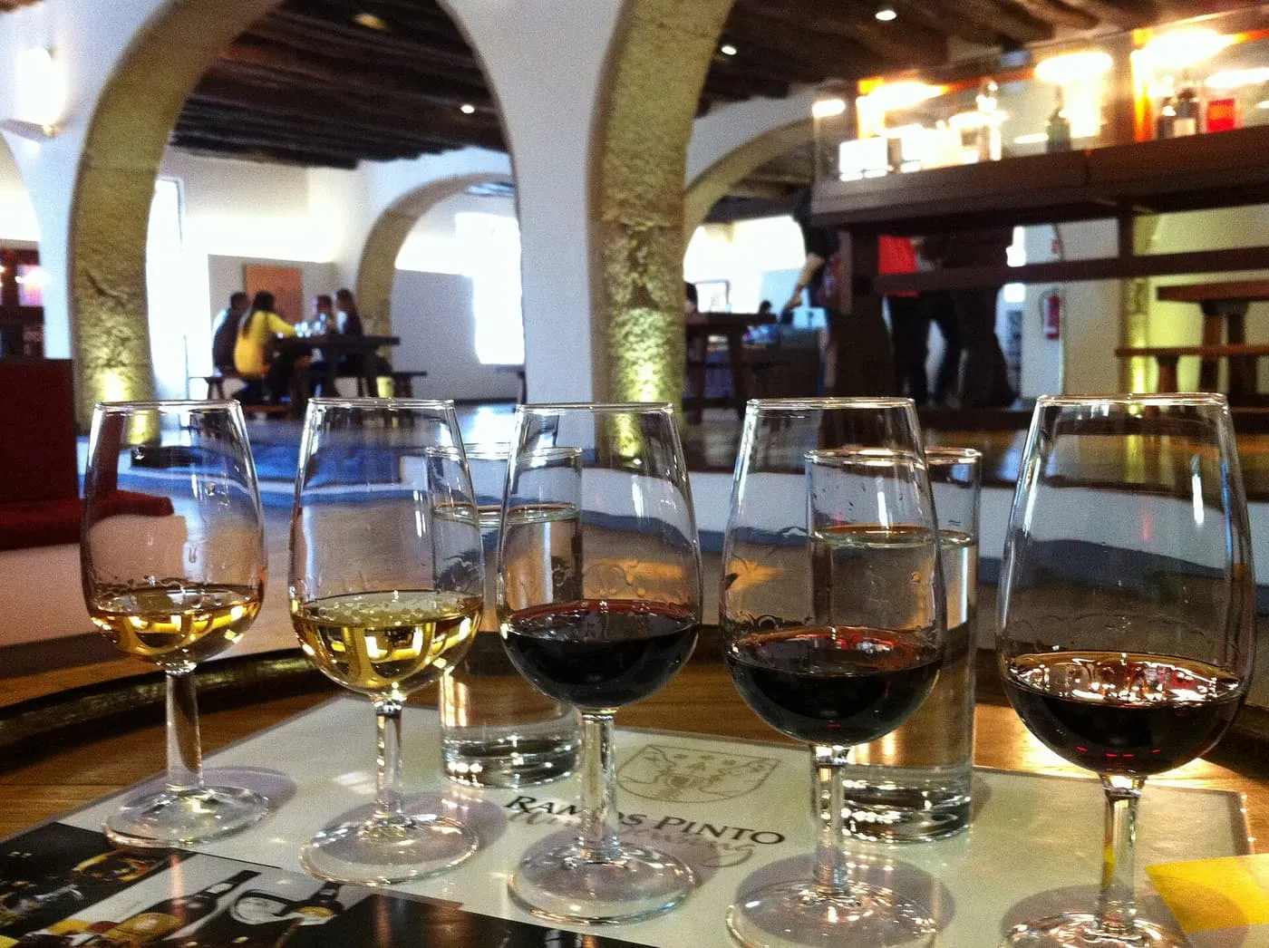 Five glasses of port, from white to ruby, set on a tasting mat in a stone-arched room