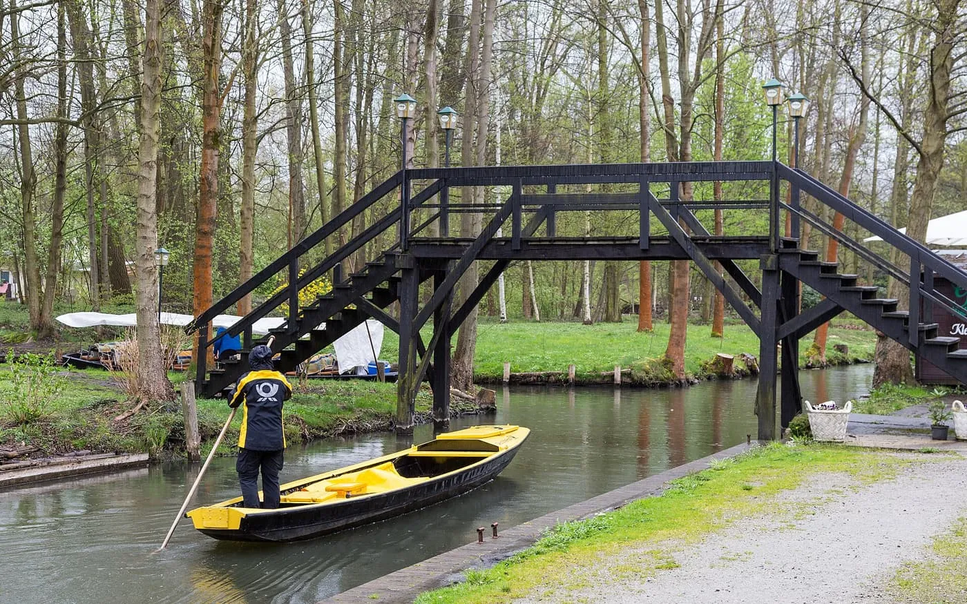 A yellow punt poled along a Spreewald canal in Lehde beneath a wooden footbridge, with trees on both banks