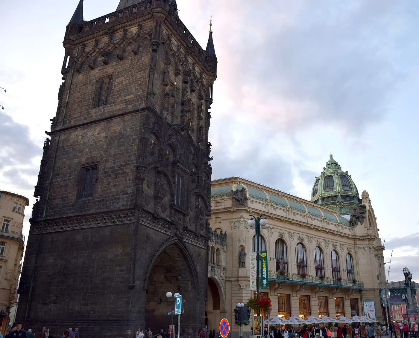 The Powder Tower, a dark Gothic tower with spires, rising beside the green dome of the Municipal House at dusk