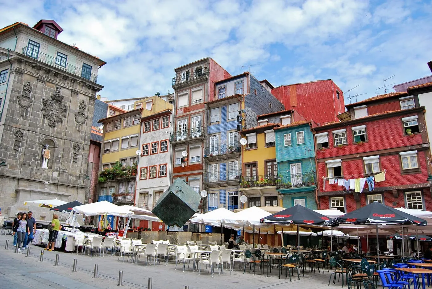 A square of tall red, yellow, blue-tiled and teal houses with café tables under parasols and a dark cube sculpture in front