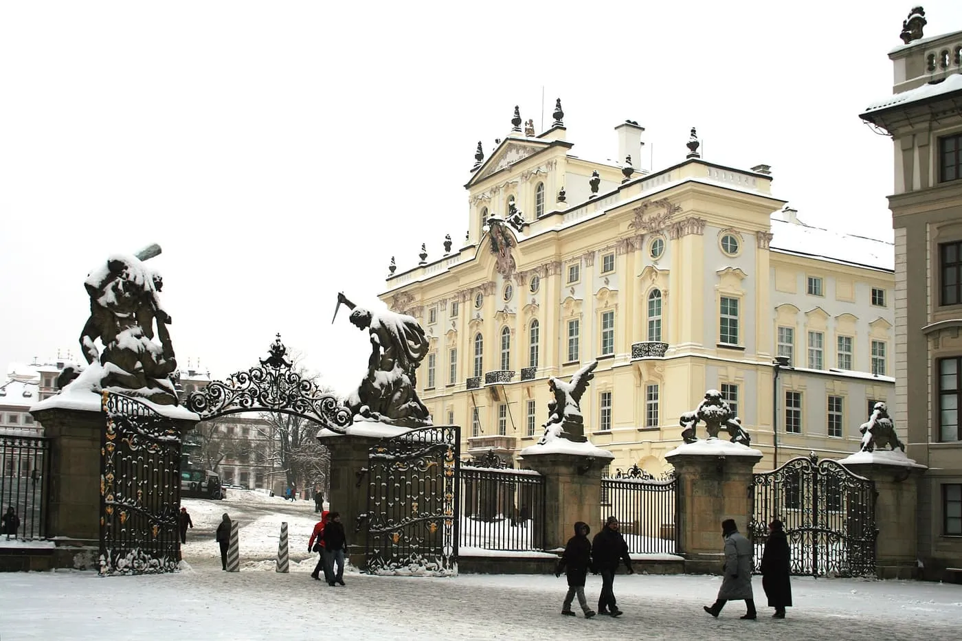 Snow on the Battling Titans gate and the yellow Baroque Archbishop's Palace at Prague Castle, with people walking across the snowy first courtyard