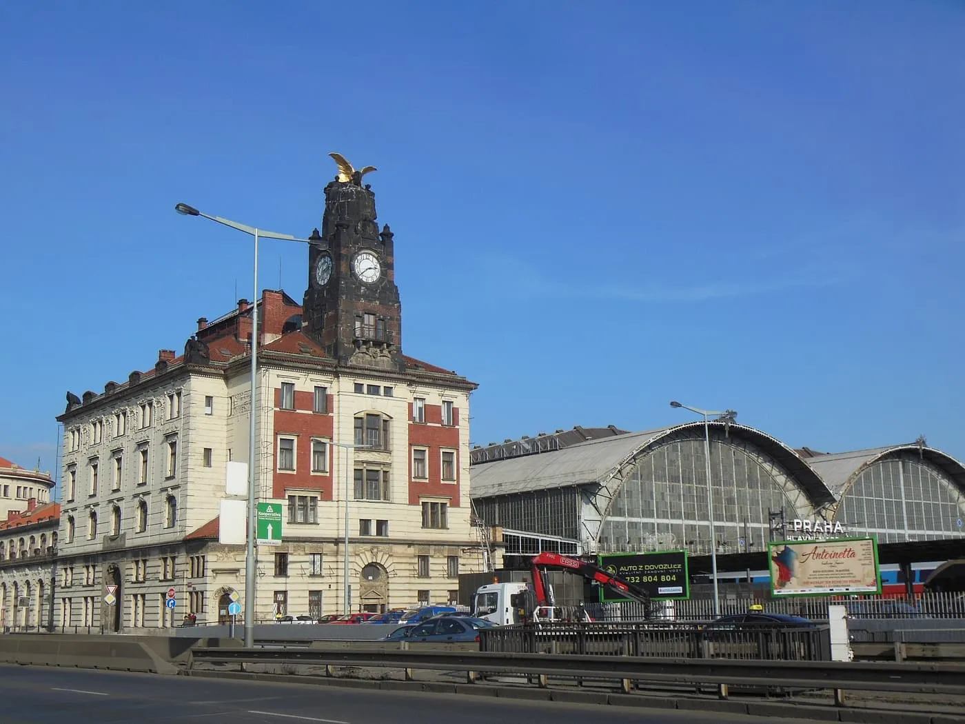 The clock tower topped by a gilded eagle beside the arched train shed of Praha hlavní nádraží under a blue sky