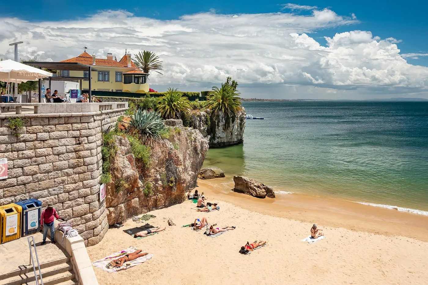 Sunbathers on the small sandy Praia da Rainha, below a stone sea wall and a yellow villa with palms above the cove