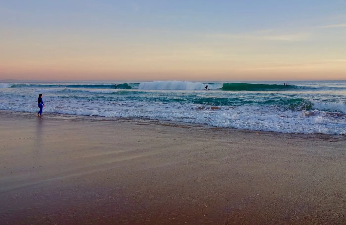 Waves breaking on a wide sandy beach at dusk, with surfers in the water