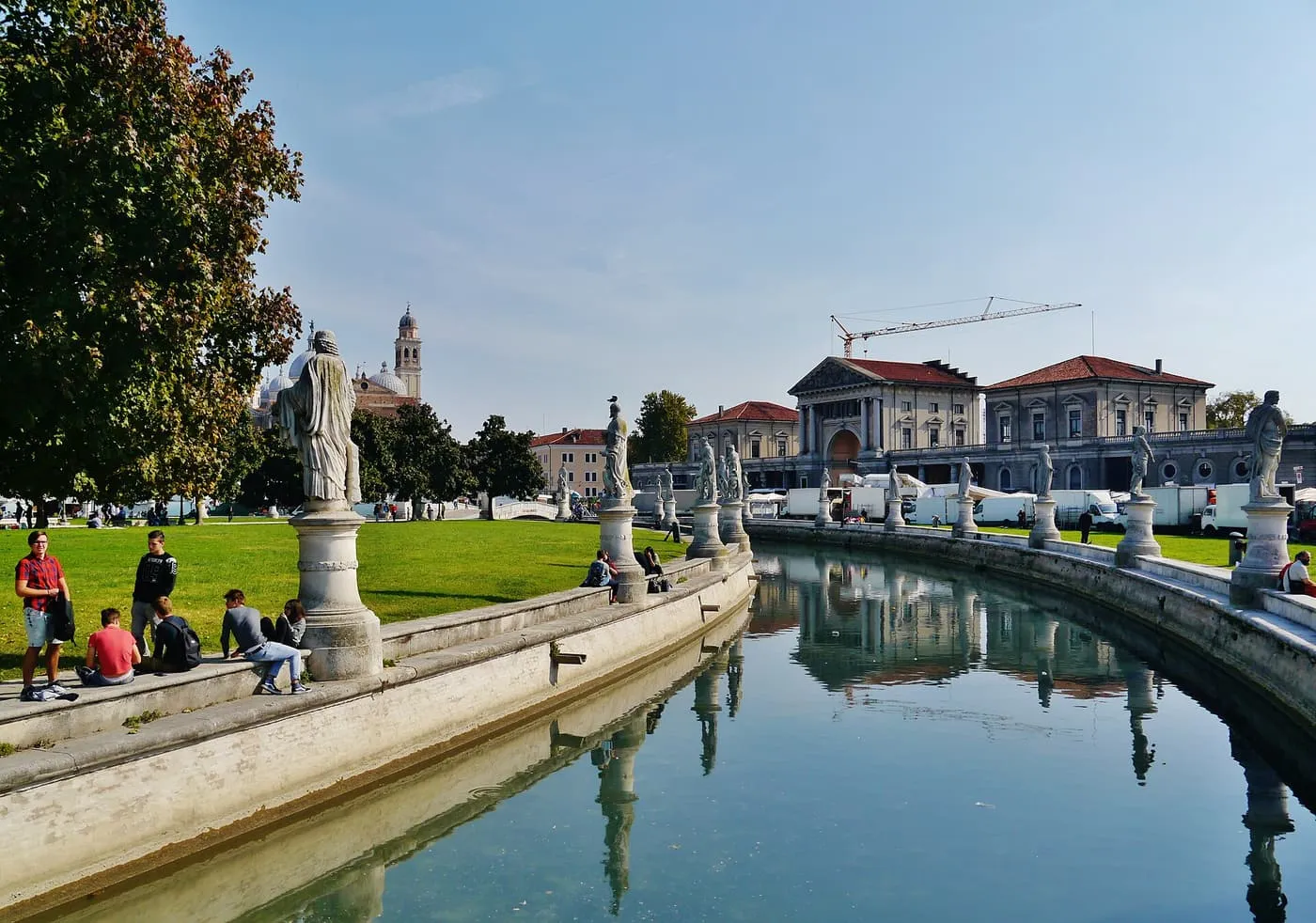 A canal ringing the green island of Prato della Valle in Padua, lined with statues on plinths, with church domes in the distance