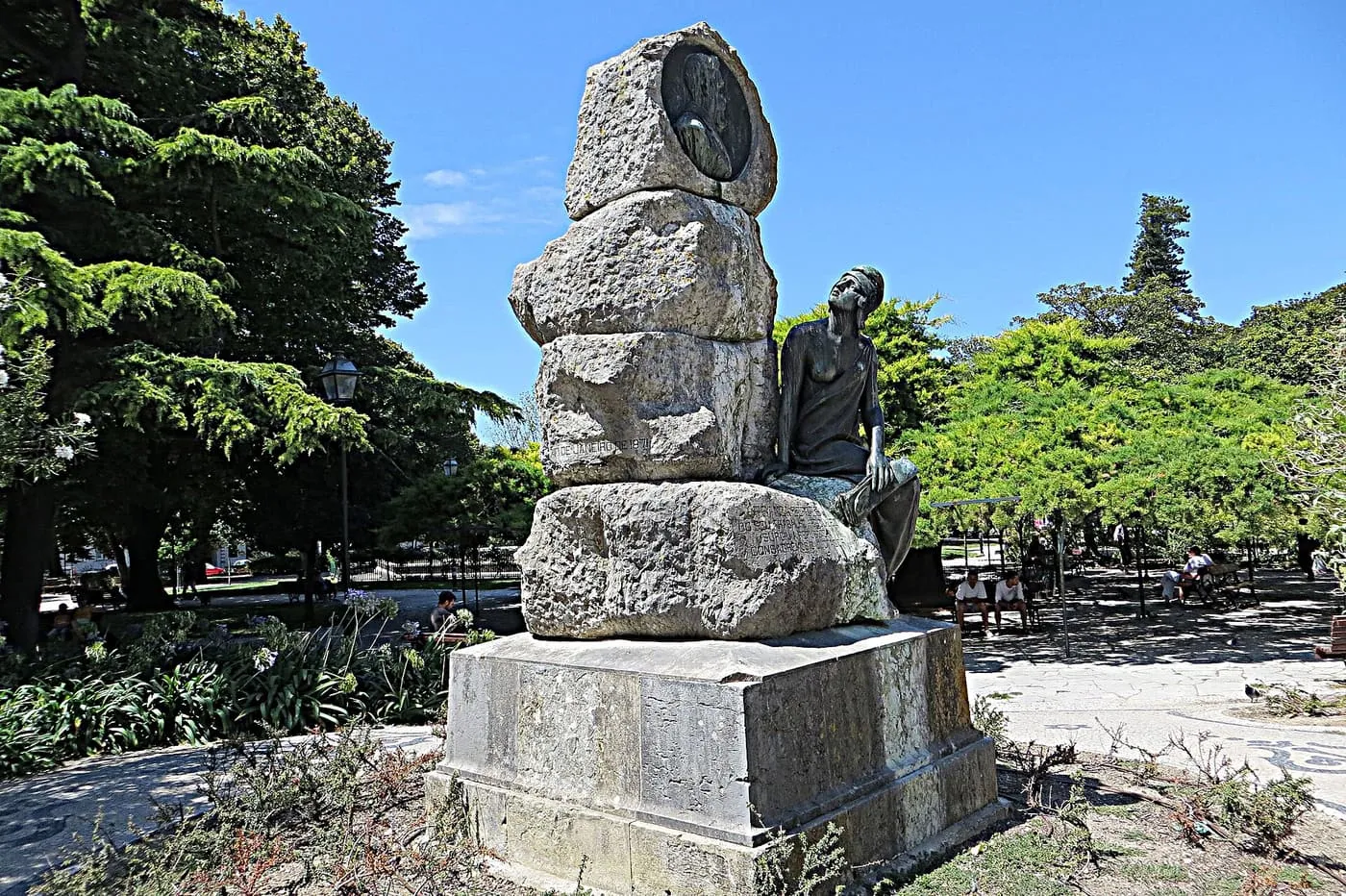 A stone monument with a bronze seated figure among trees in the Jardim do Príncipe Real