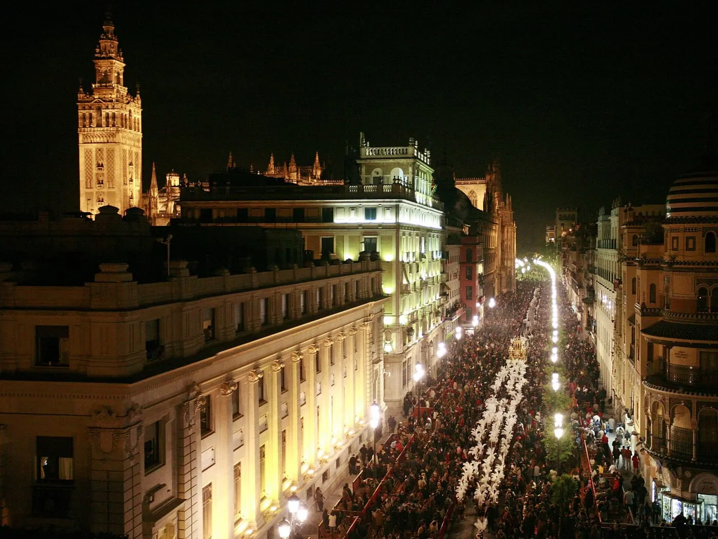 A night view from above of a Holy Week procession filling a lit street, with a float among white-plumed figures and the floodlit Giralda at the upper left