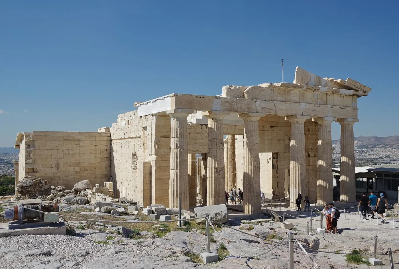The Doric columns of the Propylaia, the gateway of the Acropolis, with a few visitors beside the roped-off path under a clear blue sky