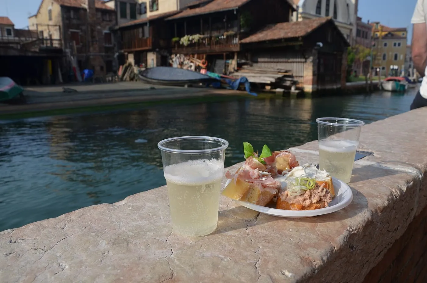Two plastic cups of Prosecco and a plate of cicchetti on a stone canal wall
