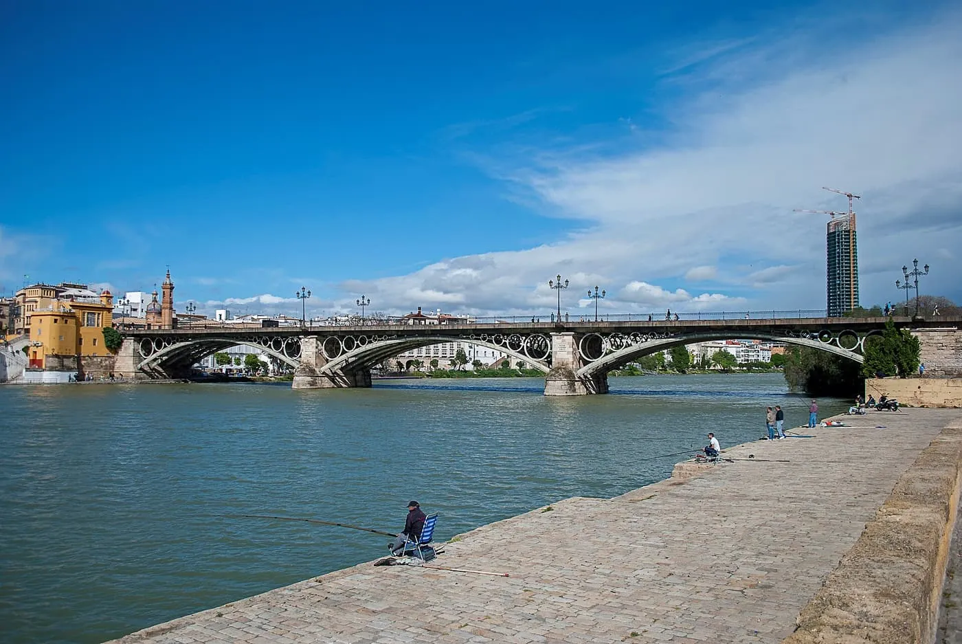 The iron arches of the Puente de Isabel II over the Guadalquivir, with anglers on the cobbled embankment and a tall tower behind