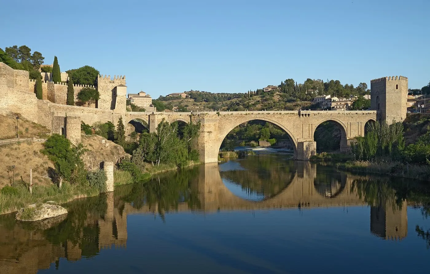 The Puente de San Martín, a medieval stone bridge with a fortified tower, reflected in the Tagus below the old town walls