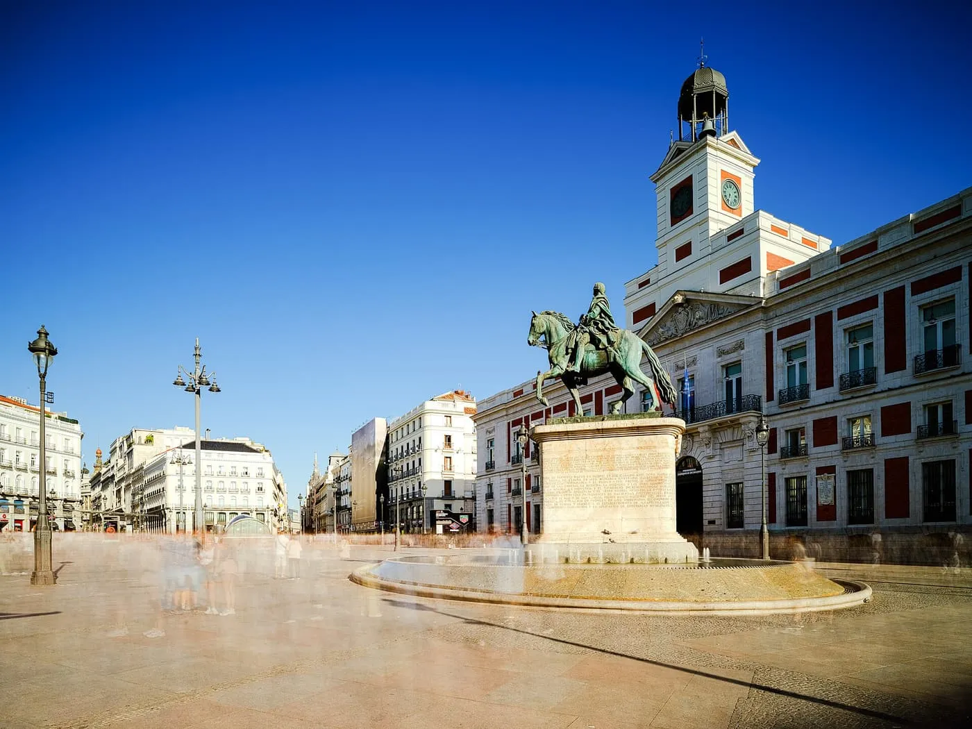 The clock tower of the Casa de Correos beside the equestrian statue of Charles III in the Puerta del Sol, under a deep blue sky, with passers-by blurred on the paving