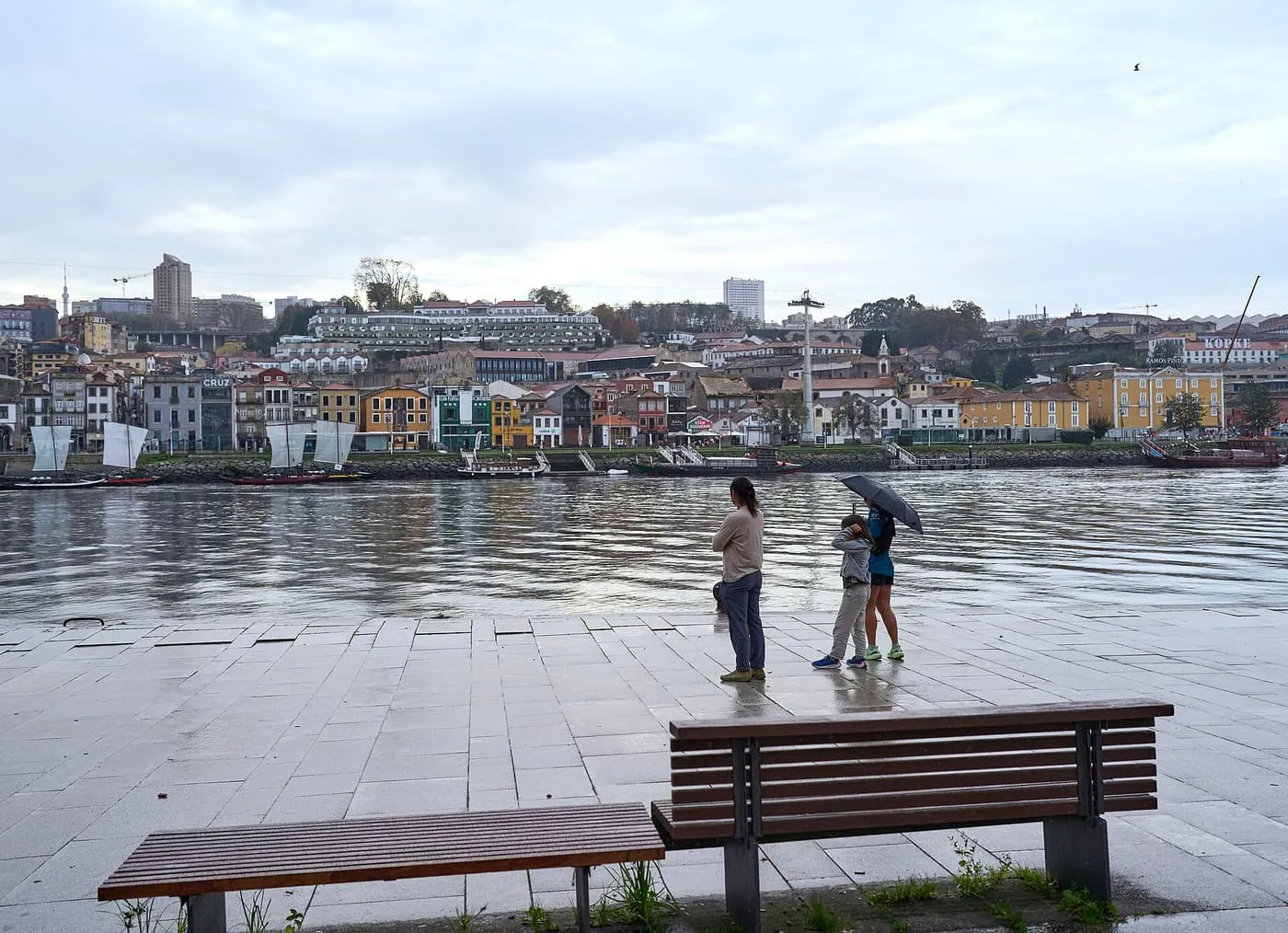 A paved riverside quay in the rain, with people under umbrellas looking across the Douro to the colourful houses on the far bank