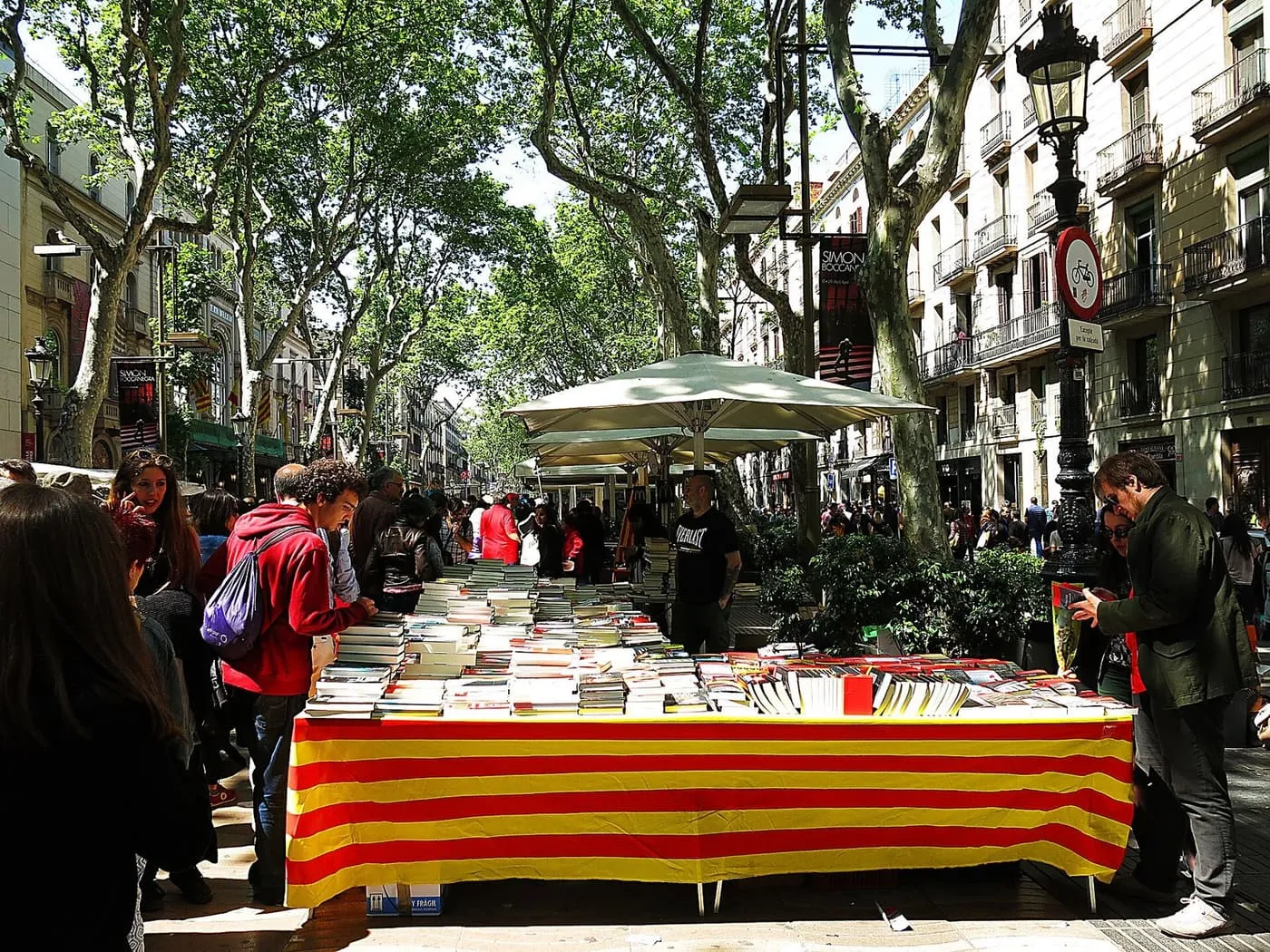 A book stall draped in the red and yellow Catalan stripes on La Rambla under plane trees, with browsers gathered round it on Sant Jordi's day