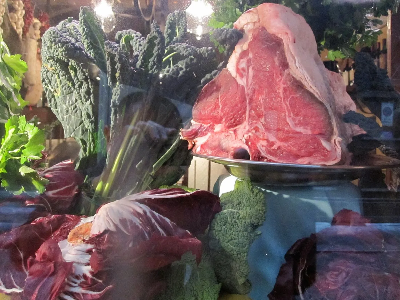 A raw T-bone fiorentina on a metal dish, surrounded by cavolo nero, radicchio and lemons in a window display