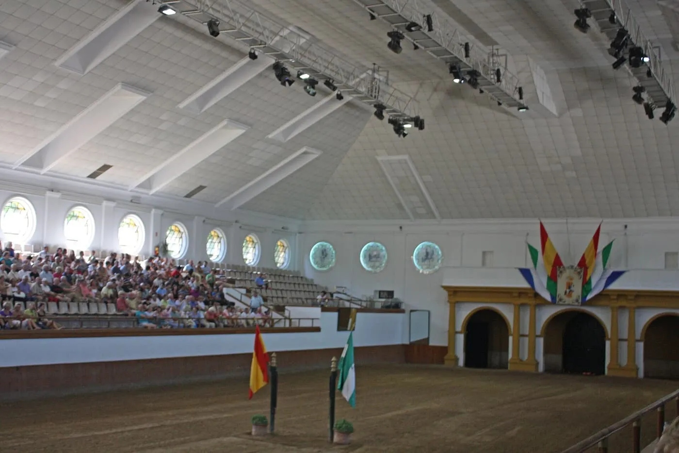 The indoor arena of the Royal Andalusian School, with raked seating on one side, flags at the centre and the stable arches at the far end