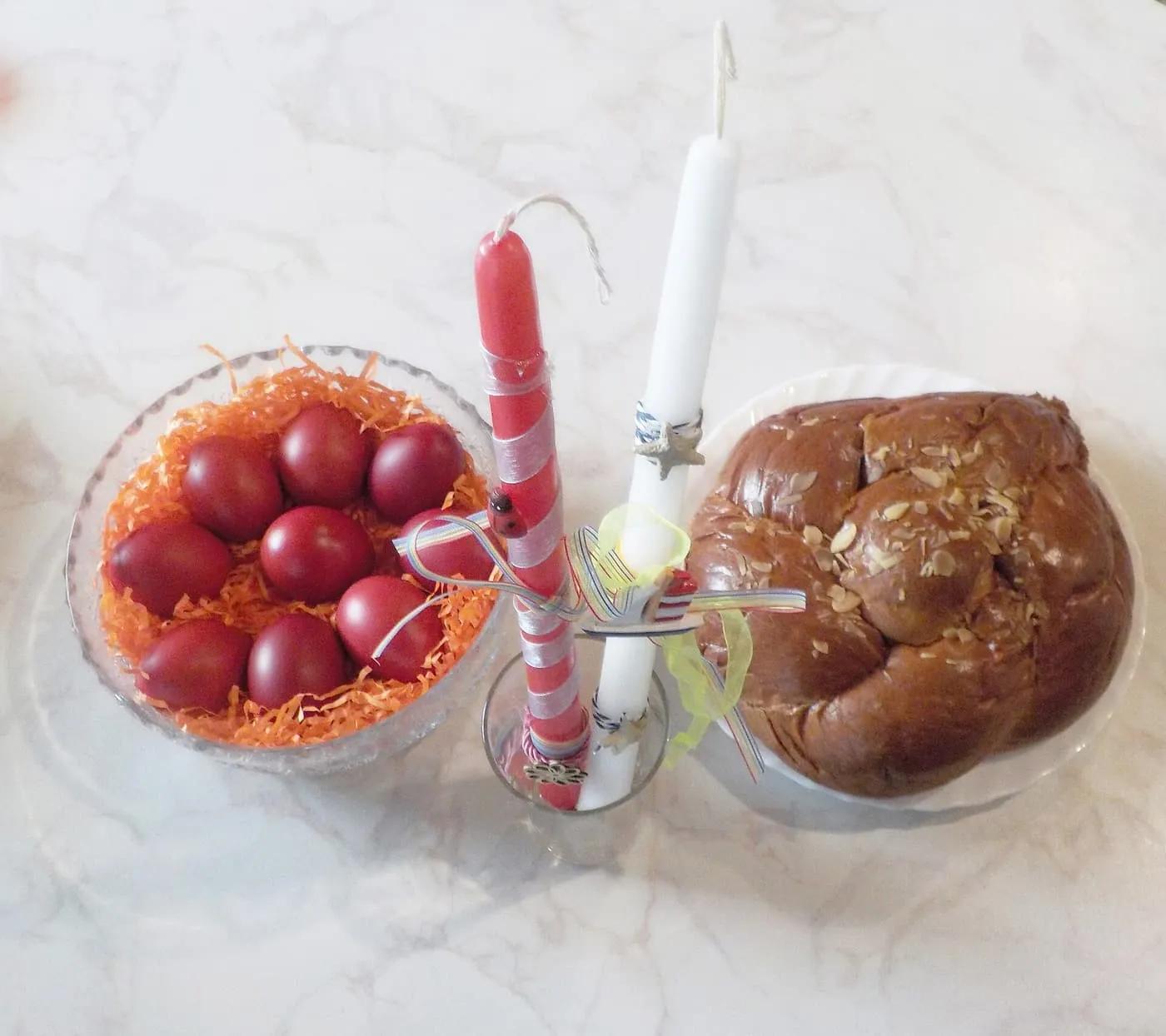 A bowl of red-dyed Easter eggs beside a red and a white Easter candle tied with ribbons and a braided tsoureki loaf topped with almonds