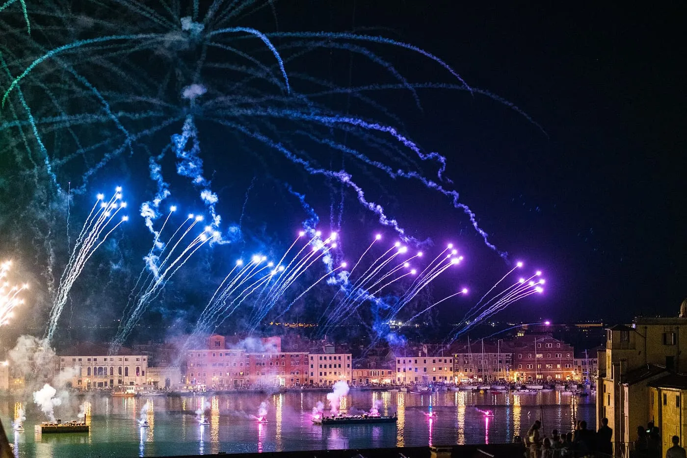 Blue and purple firework trails over a lit waterfront reflected in the water, with launch barges below