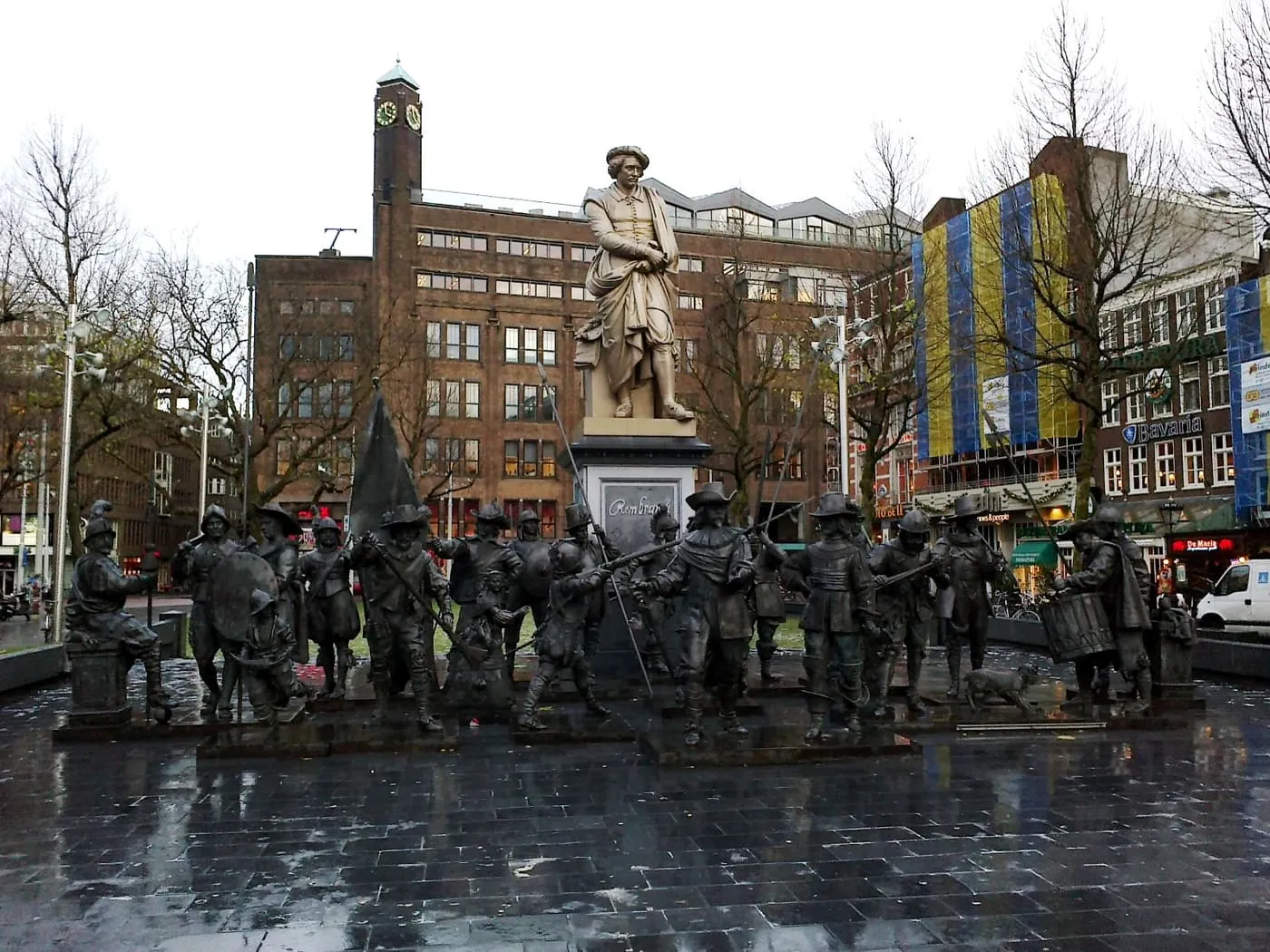 The statue of Rembrandt on its pedestal in Rembrandtplein, with a group of bronze figures from The Night Watch round its base on wet paving
