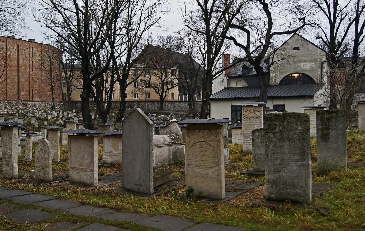 Weathered stone gravestones in the Remuh cemetery among bare trees, with the synagogue roof behind