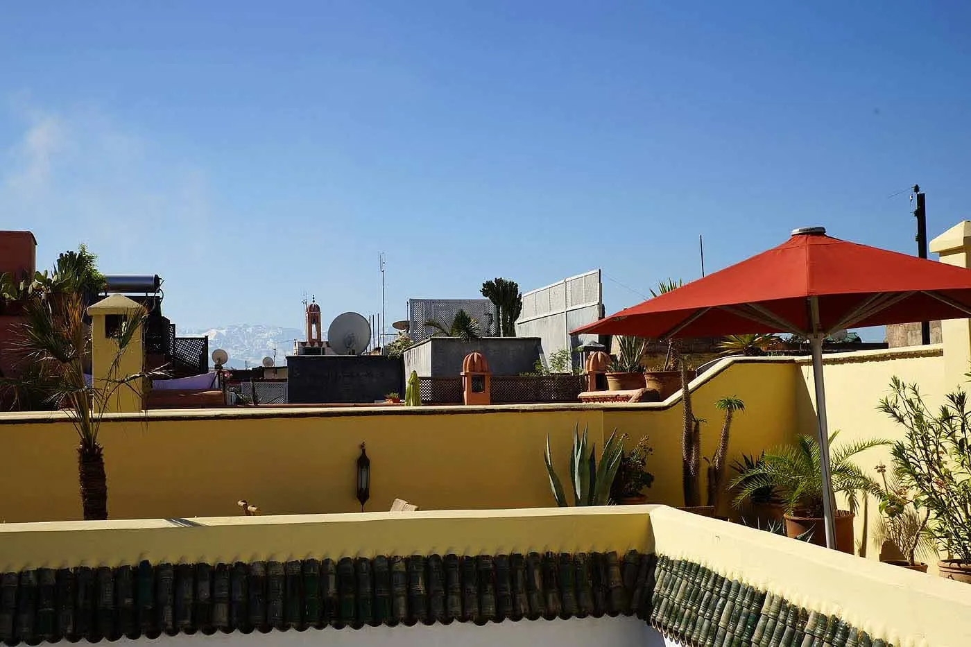 A yellow-walled roof terrace with a red parasol and potted palms, and the snow-topped Atlas faint on the horizon
