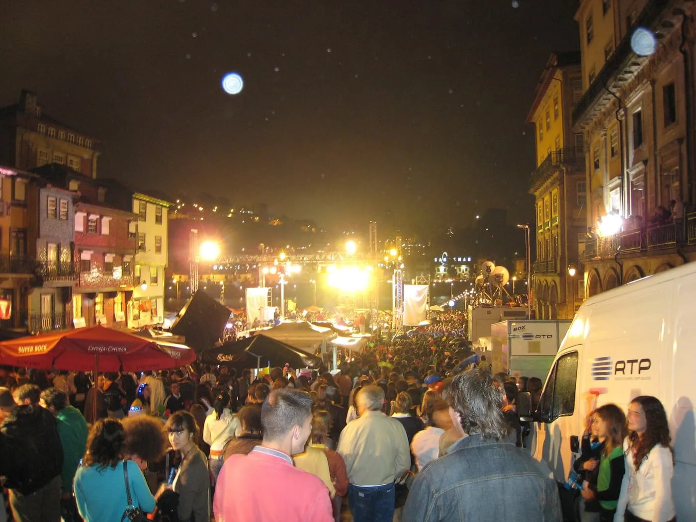 A crowd packed into a lit street of old houses at night, with a stage and a television van at the back