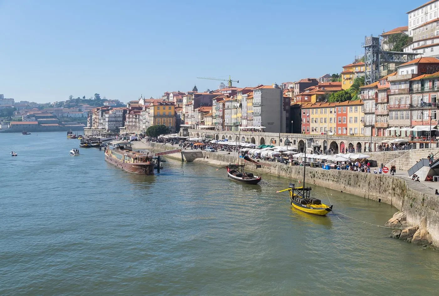 Porto's Ribeira quayside seen from the Dom Luís I bridge, with café umbrellas and two rabelo boats on the Douro