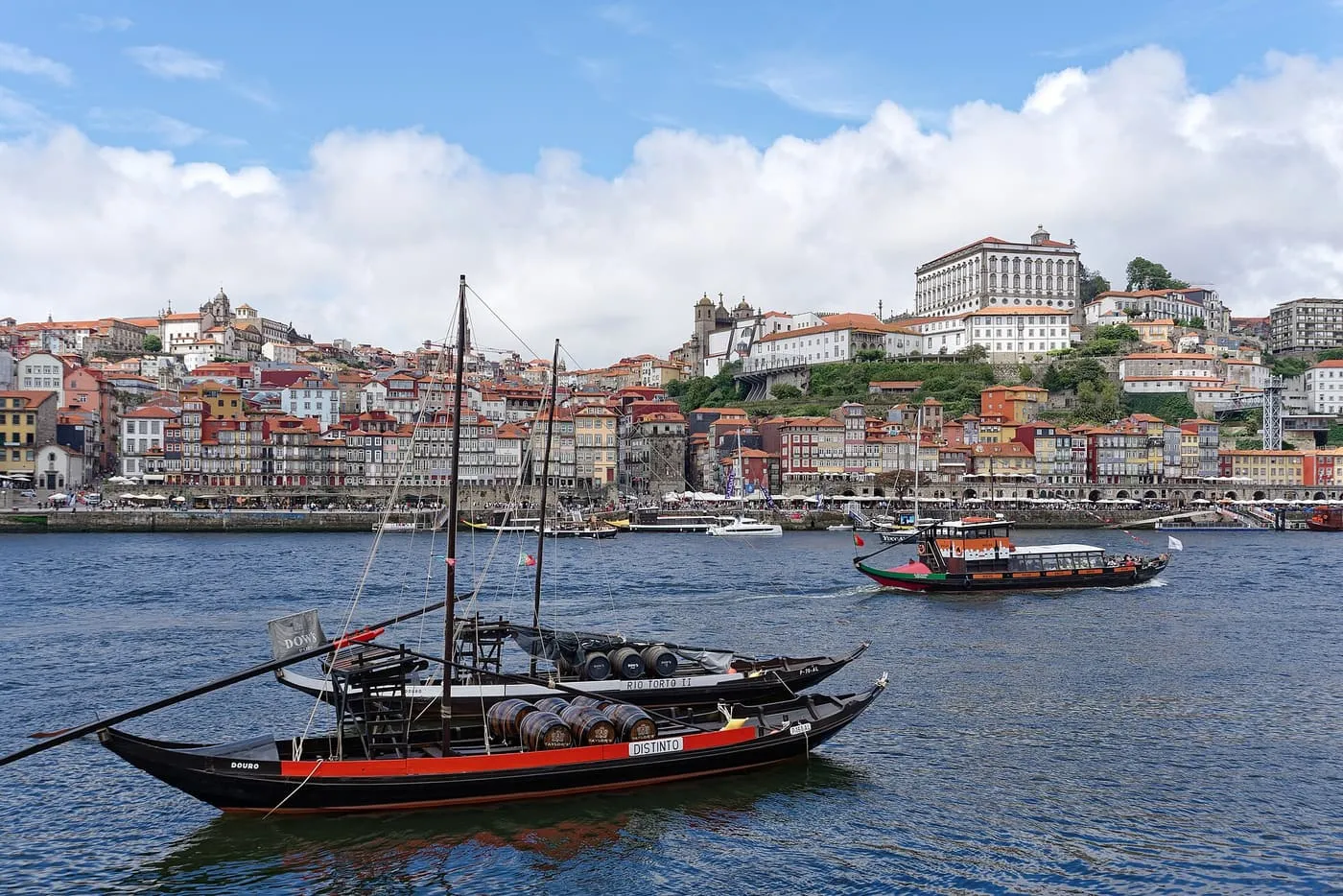 Two rabelo boats loaded with port barrels on the Douro, with Porto's Ribeira houses climbing the hill behind