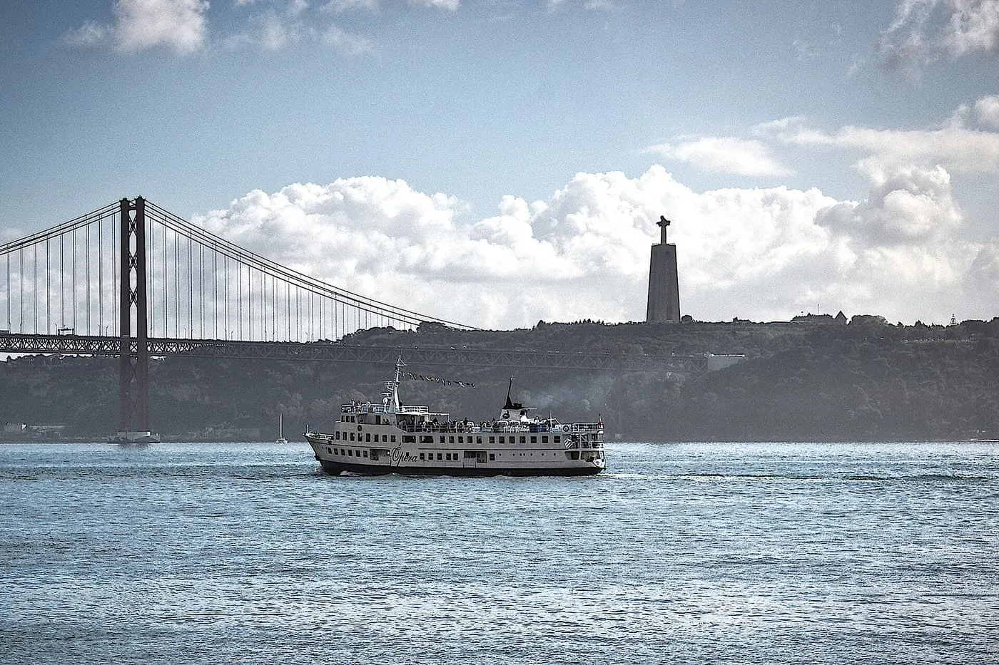 A white river cruise boat on the Tagus, with the 25 de Abril Bridge and the Cristo Rei statue behind
