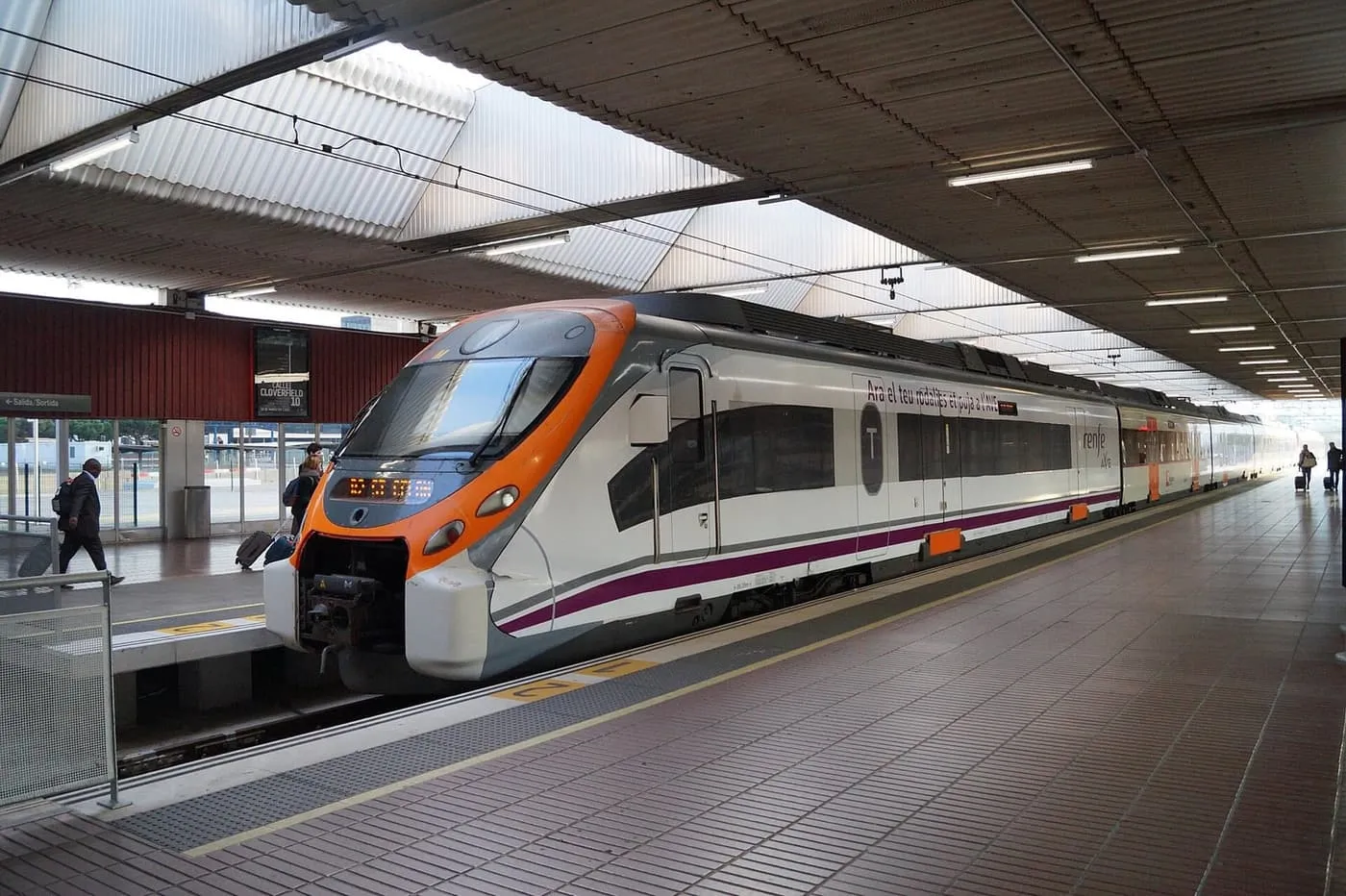 A white and orange Rodalies train waiting under the canopy at Aeroport station