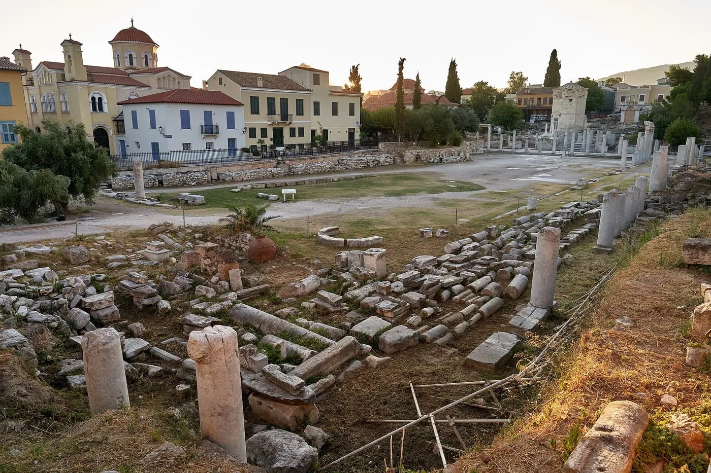 Fallen column drums and stumps of columns in the open court of the Roman Agora, with older houses and a church behind and the Tower of the Winds in the distance at the right