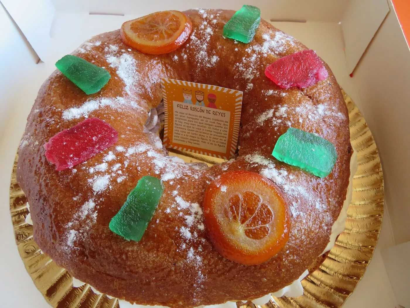 A roscón de Reyes seen from above, a ring of sweet bread topped with candied orange slices, red and green candied fruit and icing sugar, on a gold card in a box