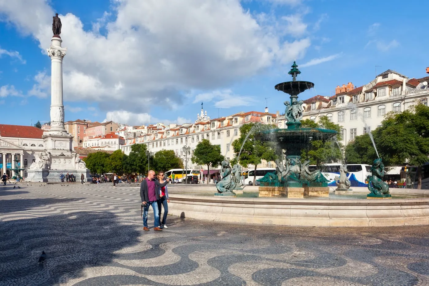 Rossio's wave-patterned pavement, a bronze fountain, the column of Pedro IV and the buildings round the square under a blue sky with clouds