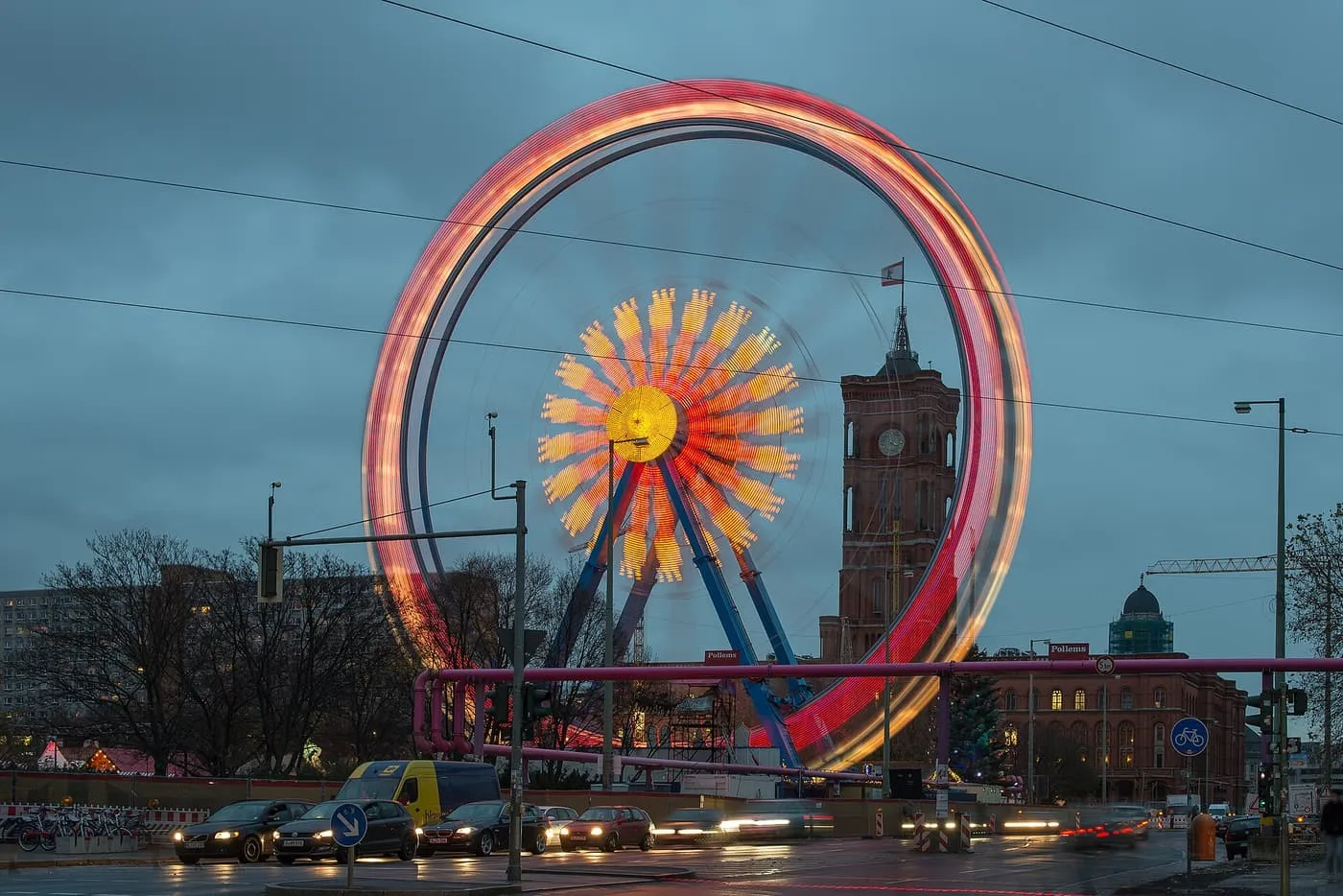 A Ferris wheel spinning in a blur of red and orange light in front of the brick tower of the Rotes Rathaus at dusk