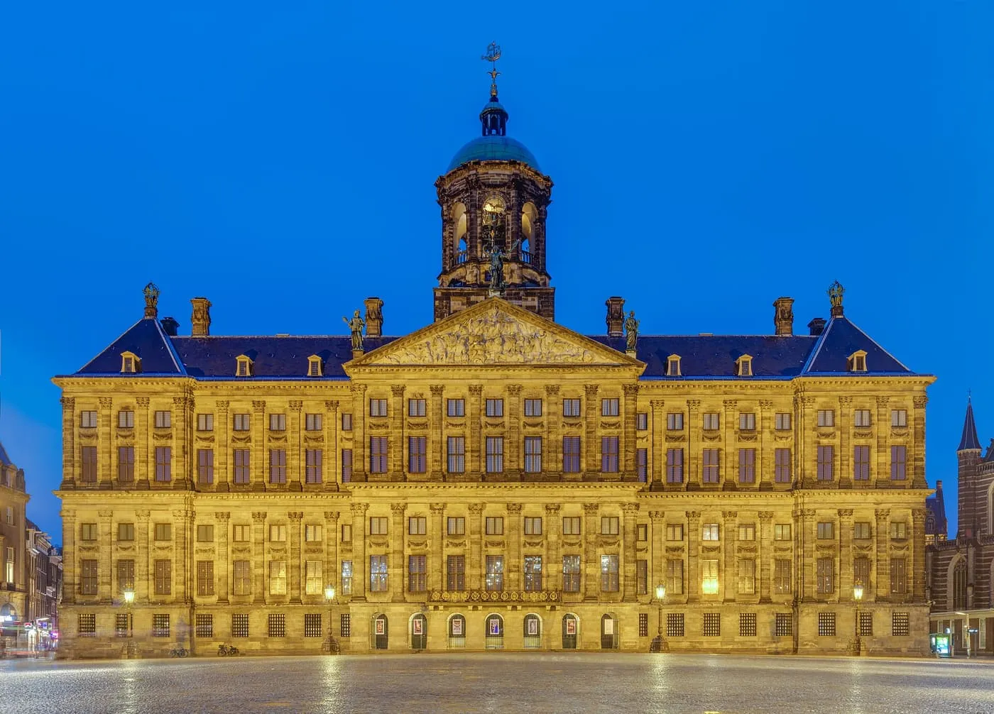 The Royal Palace on Dam Square at dusk, a long pale stone building lit gold, with a central pediment and a domed cupola