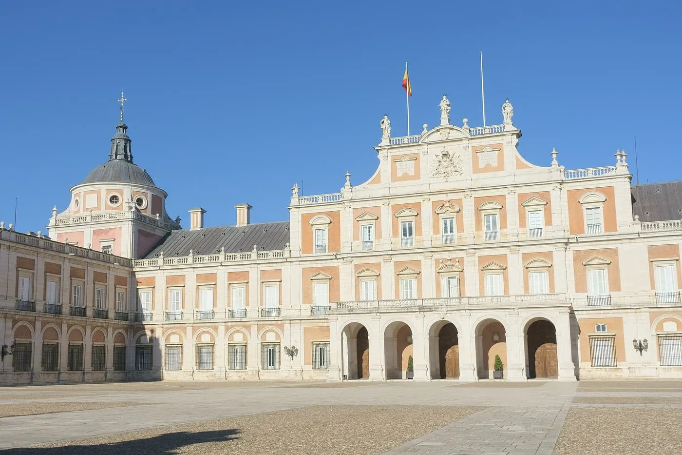 The pink brick and white stone west façade of the Royal Palace of Aranjuez, with a domed corner pavilion and a statue-topped central block, across an empty gravel courtyard