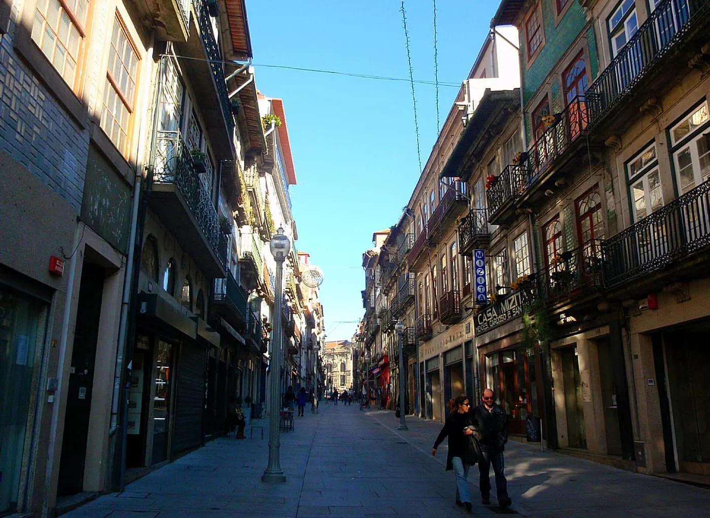 A narrow pedestrian street lined with balconied buildings, with a few people walking and a blue sky above