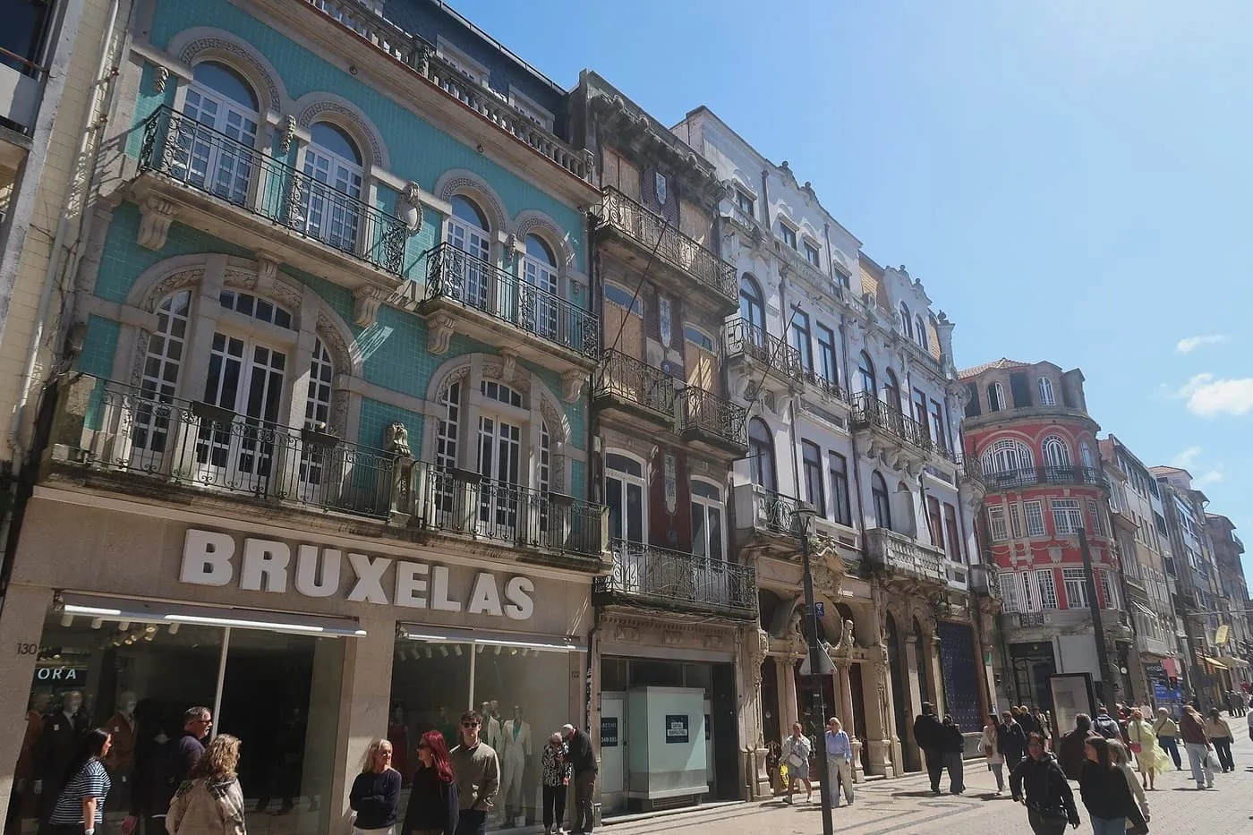 A busy pedestrian street with a blue-tiled balconied corner building, shop fronts and crowds of walkers under a blue sky