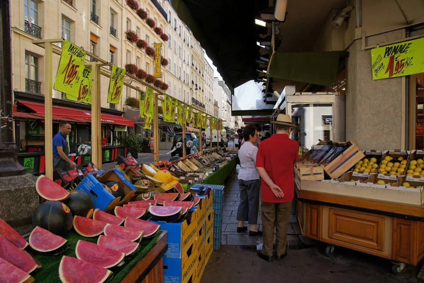 Fruit stalls with watermelons and apples along the pedestrian Rue Cler, under apartment buildings with flower boxes